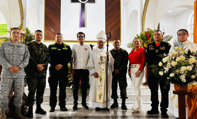 Agentes de la Policía, soldados y personal de la Fuerza Aérea junto a dos pastores con vestimenta blanca. Varias personas con camisas de distintos colores están de pie dentro de una iglesia decorada con flores blancas.
