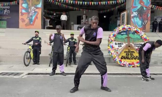 Presentaci&oacute;n art&iacute;stica en un espacio p&uacute;blico de Medell&iacute;n: varias personas realizan una danza frente a un escenario, mientras integrantes de la Polic&iacute;a Nacional en bicicleta observan al fondo. Se aprecia decoraci&oacute;n colorida y un c&iacute;rculo floral con un mensaje alusivo al di&aacute;logo.