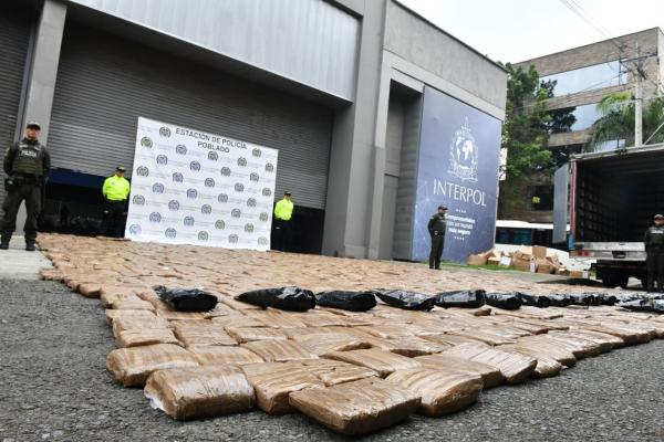 Se observa una gran cantidad de paquetes rectangulares envueltos en pl&aacute;stico, en el suelo como resultado de una incautaci&oacute;n de marihuana. Al fondo se ven uniformados de la Polic&iacute;a Nacional, un cami&oacute;n y un banner institucional.   